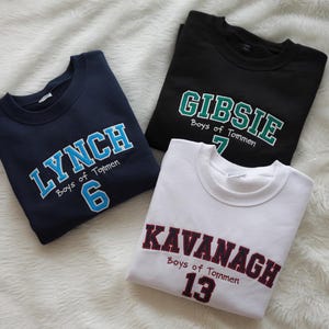May include: Three navy blue, black, and white sweatshirts with embroidered text. The navy blue sweatshirt says "LYNCH Boys of Tommen 6", the black sweatshirt says "GIBSIE Boys of Tommen 7", and the white sweatshirt says "KAVANAGH Boys of Tommen 13".