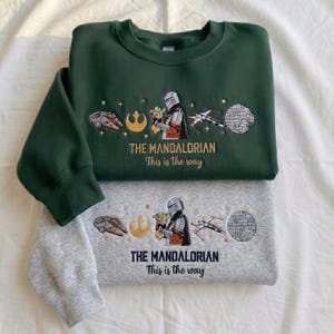 May include: A dark green sweatshirt and a light gray sweatshirt, each featuring embroidered Star Wars designs including a Mandalorian character, a small green creature, a spaceship, and the text "THE MANDALORIAN This is the way" in gold or black.