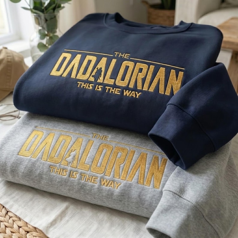 Może przedstawiać: Dwie bluzy, jedna granatowa i jedna szara, ułożone jedna na drugiej. Obie mają złoty napis "THE DADALORIAN THIS IS THE WAY". Rękaw granatowej bluzy jest złożony na szarej.