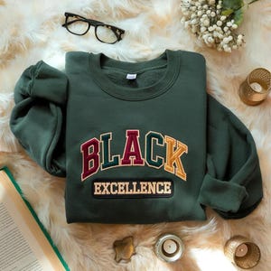 Könnte beinhalten: Ein dunkelgrünes Langarm-Sweatshirt mit dem gestickten Text BLACK EXCELLENCE in einer College-Schriftart. Das Wort BLACK ist in Bordeaux, Dunkelgrün und Gold gebogen, während EXCELLENCE auf einem rechteckigen Aufnäher in Creme steht.