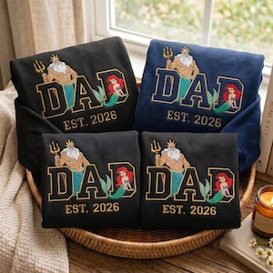 May include: Four folded sweatshirts in black and navy blue featuring embroidered graphics of King Triton and Ariel from The Little Mermaid. The design includes the text DAD in large block letters and EST. 2026 in smaller gold-coloured lettering.