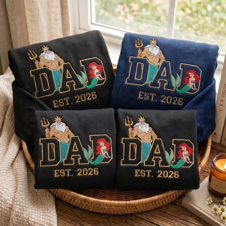 May include: Four folded sweatshirts in black and navy blue featuring embroidered graphics of King Triton and Ariel from The Little Mermaid. The design includes the text DAD in large block letters and EST. 2026 in smaller gold-coloured lettering.