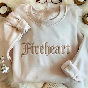 Puede incluir: Una sudadera beige claro con la palabra "Fireheart" bordada en una fuente gótica beige más oscura. La sudadera tiene cuello redondo y puños. La imagen está tomada desde arriba.