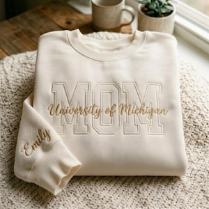 May include: Cream-colored sweatshirt with the word "MOM" embroidered in a block letter style, with "University of Michigan" below. The sleeve cuff has the name "Emily" embroidered in a script font. The sweatshirt is folded on a textured surface.
