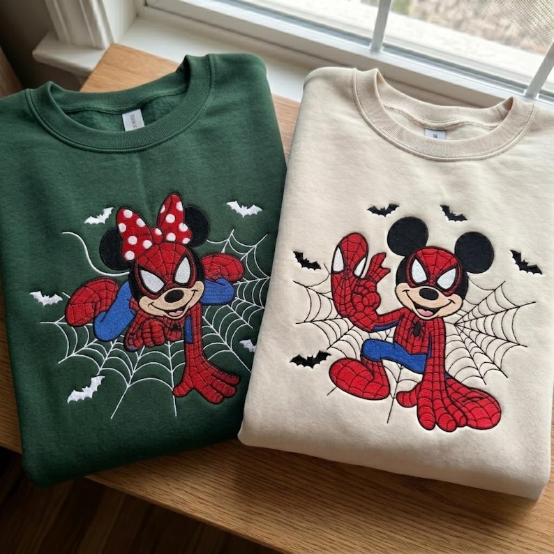 May include: A dark green sweatshirt and a beige sweatshirt, each featuring an embroidered graphic of a cartoon mouse dressed in a red and blue spider-themed costume. Both designs include a white spiderweb background and small black bat silhouettes.