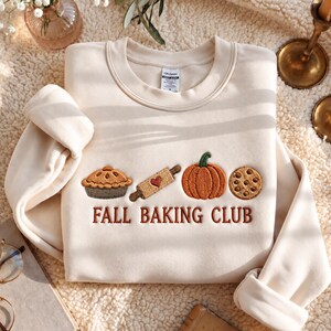 May include: A cream-colored crewneck sweatshirt featuring embroidered icons of a pie, a rolling pin with a heart, a pumpkin, and a chocolate chip cookie. Below the icons, the text FALL BAKING CLUB is embroidered in brown thread.