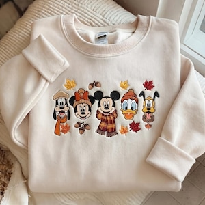May include: A cream-coloured crewneck sweatshirt featuring an embroidered design of five cartoon characters wearing autumn accessories. The characters are surrounded by small embroidered maple leaves and acorns in shades of orange, red, and brown.