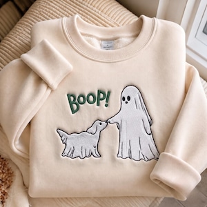 Peut inclure: Un sweat-shirt à col rond de couleur crème avec une broderie représentant un fantôme blanc touchant le nez d'un chien blanc. Le mot BOOP! est brodé en vert foncé au-dessus des personnages. Le vêtement possède un col et des poignets côtelés.