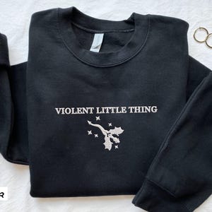 Puede incluir: Sudadera negra con un dise&ntilde;o bordado en blanco que dice "VIOLENT LITTLE THING" y una peque&ntilde;a imagen de un drag&oacute;n con acebo.