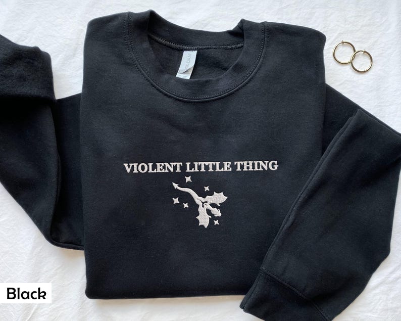 Puede incluir: Sudadera negra con un dise&ntilde;o bordado en blanco que dice "VIOLENT LITTLE THING" y una peque&ntilde;a imagen de un drag&oacute;n con acebo.