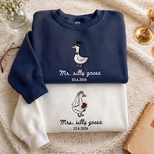 May include: Two sweatshirts, one navy blue and one white, featuring embroidered goose designs. The navy sweatshirt says "Mr. silly goose" and the white one says "Mrs. silly goose", both with a date. The goose on the navy sweatshirt wears a top hat.