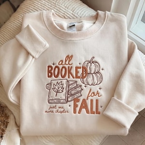 May include: A cream-colored crewneck sweatshirt featuring brown embroidery. The design includes a stack of books, a pumpkin, and the text "all BOOKED for FALL" and "just one more chapter" with small star accents surrounding the central graphic.