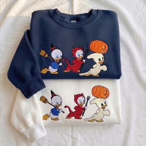 May include: A navy blue sweatshirt and a white sweatshirt, each featuring an embroidered graphic of three cartoon ducks in Halloween costumes: one as a witch with a broom, one as a red devil with a pitchfork, and one as a ghost balancing a pumpkin.