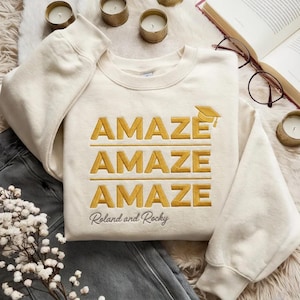 May include: Cream-colored sweatshirt with the word "AMAZE" embroidered in gold three times, topped with a graduation cap graphic. The text "Roland and Rocky" is embroidered below. The sweatshirt is laid on a white fluffy surface with jeans and candles.