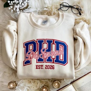 May include: Cream-colored sweatshirt with the text "PHD Dr. Sarah EST. 2026" in blue and pink lettering. The sweatshirt has a crew neck and long sleeves. The design is embroidered with a collegiate style font.