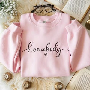 May include: A light pink sweatshirt with the word "homebody" in black script and a small heart. The sweatshirt is folded on a fluffy white surface, with reading glasses and a book in the background.