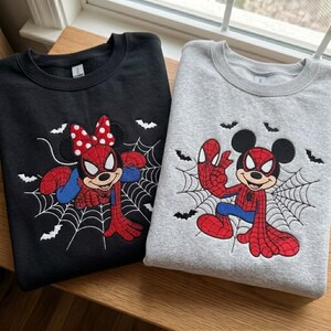 May include: A black sweatshirt and a gray sweatshirt, each featuring an embroidered graphic of a cartoon mouse dressed as a superhero with a spiderweb background and small black bats. The black sweatshirt features a mouse with a red polka dot bow.