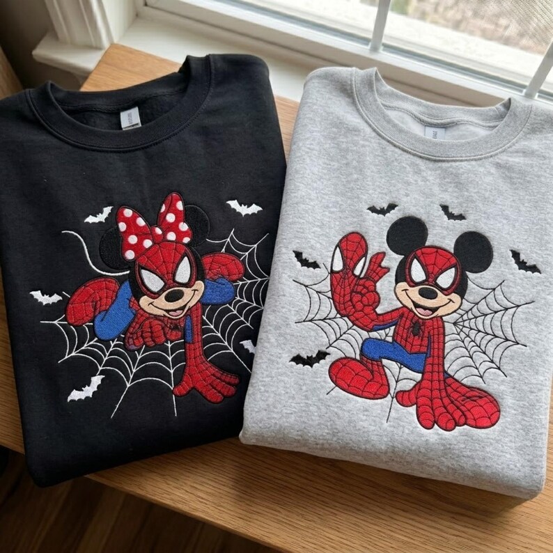 May include: A black sweatshirt and a gray sweatshirt, each featuring an embroidered graphic of a cartoon mouse dressed as a superhero with a spiderweb background and small black bats. The black sweatshirt features a mouse with a red polka dot bow.