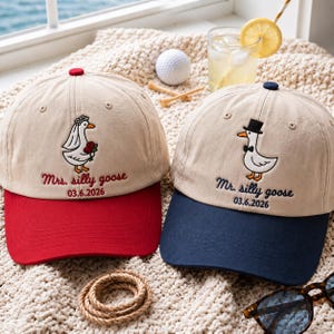 May include: Two beige baseball caps featuring embroidered goose designs. The left cap has a red brim, a goose wearing a veil and holding red flowers, with the text Mrs. silly goose 03.6.2026. The right cap has a navy brim, a goose in a top hat, and Mr. silly goose 03.6.2026.