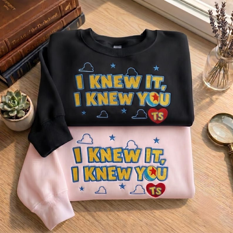 May include: A black sweatshirt and a light pink sweatshirt, each featuring embroidered text that reads I KNEW IT, I KNEW YOU with a colorful ball icon, a red heart with the letters TS, and small blue cloud and star shapes across the front.
