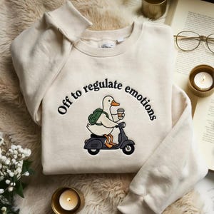May include: A cream-coloured crewneck sweatshirt featuring an embroidered graphic of a white duck wearing a green backpack and holding a coffee cup while riding a dark grey scooter. The text "Off to regulate emotions" is arched above the duck.