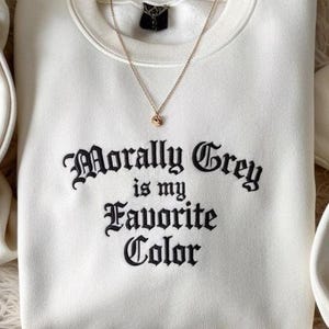 May include: A white sweatshirt with the embroidered text "Morally Grey is my Favorite Color" in black gothic lettering. A gold necklace with a small pendant rests on the sweatshirt.