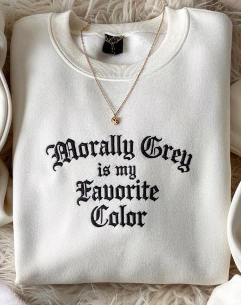 May include: A white sweatshirt with the embroidered text "Morally Grey is my Favorite Color" in black gothic lettering. A gold necklace with a small pendant rests on the sweatshirt.