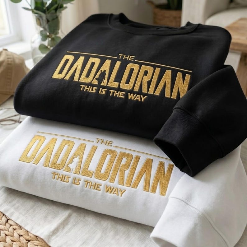 Może przedstawiać: Dwie bluzy, jedna czarna i jedna biała, są ułożone jedna na drugiej. Obie mają złoty napis "THE DADALORIAN" z sylwetką i "THIS IS THE WAY". Widoczny jest rękaw czarnej bluzy. Bluzy są złożone na białej powierzchni.