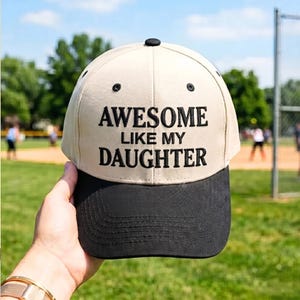 Puede incluir: Gorra de béisbol beige y negra con el texto "AWESOME LIKE MY DAUGHTER" bordado en negro. La gorra tiene una visera curva y un botón en la parte superior. El fondo muestra un campo de béisbol y un día soleado.