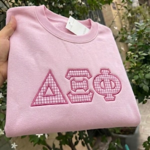 Peut inclure: Un sweat-shirt à col rond rose clair orné de trois lettres grecques brodées dans un motif vichy rose et blanc. Les lettres sont soulignées d'un fil rose uni et une étiquette rectangulaire blanche est fixée à l'intérieur du col.