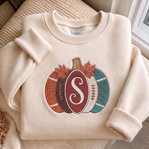 Pode incluir: Uma camisola de gola redonda cor de creme com um desenho de abóbora bordado na parte da frente. A abóbora está dividida em secções de cor laranja, castanho, creme e azul-petróleo, com uma letra S grande e duas folhas de ácer.