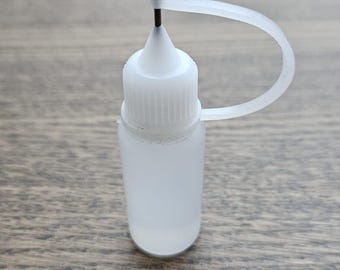 May include: A small, translucent white plastic bottle with a black needle tip and a white cap. The bottle has a curved, attached white plastic loop. The bottle is sitting on a wooden surface.