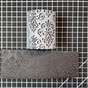 May include: A close-up of a gray, textured, cylindrical stamp with a damask pattern. The stamp is sitting on a black, textured, rectangular surface.