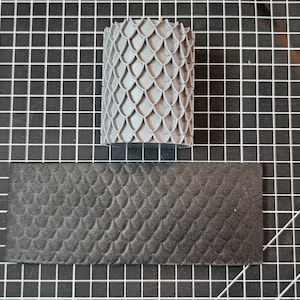 May include: A gray cylindrical stamp with a scale pattern sits above a rectangular piece of black material with the same scale pattern. The objects are on a black and white grid surface. The stamp is likely used for leather or other craft projects.