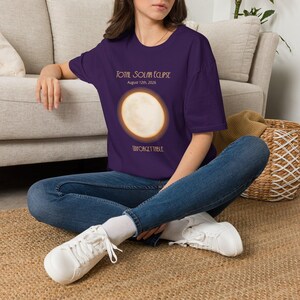 Può includere: Una t-shirt viola scuro con una grafica di un sole splendente durante un'eclissi solare totale. Il testo recita Total Solar Eclipse, August 12th, 2026, e Unforgettable. La maglietta ha maniche corte, girocollo e viene indossata con jeans blu.