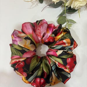May include: A black satin scrunchie with a floral print featuring pink, yellow, and red flowers, green leaves, and red cherries.