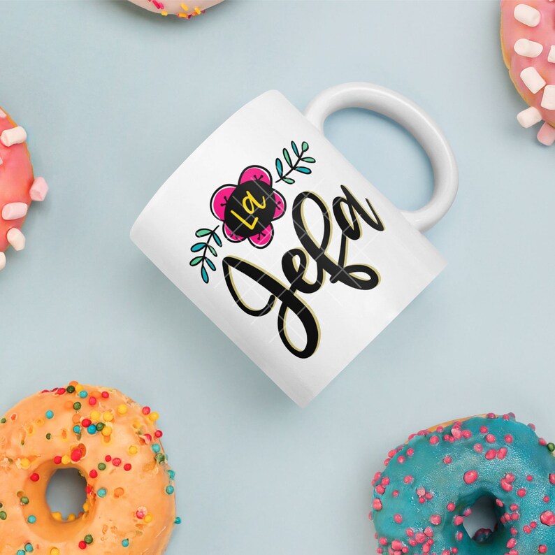 May include: White ceramic mug with a black and gold design that says "La Jefa" with a pink flower and green leaves.