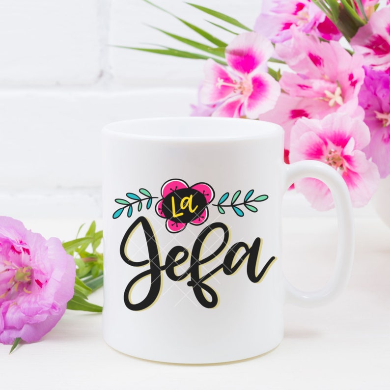 May include: White ceramic mug with a black and white floral design and the text "La Jefa" in black script. The flower is pink, yellow, and blue.