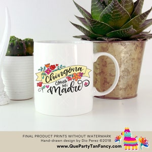 May include: White ceramic mug with a colorful floral design and the text "Chingona como mi madre" in black script. The mug has a handle and is sitting on a white surface.