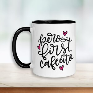 May include: White ceramic coffee mug with a black rim. The mug has a black handwritten message that says "pero first cafecito" with pink hearts.