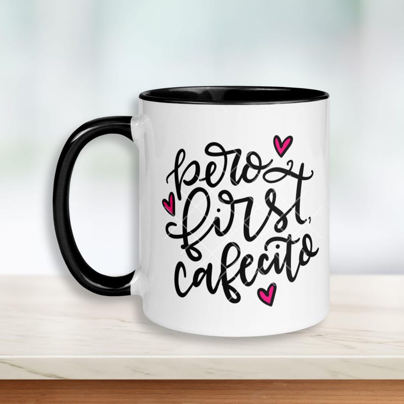 May include: White ceramic coffee mug with a black rim. The mug has a black handwritten message that says "pero first cafecito" with pink hearts.