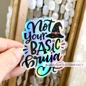May include: A holographic sticker with the text "Not your basic bruja" in black lettering. A black witch hat with a gold buckle and stars is above the text. The sticker has a rainbow iridescent background.