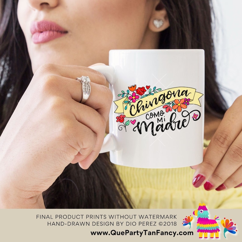 May include: White ceramic mug with a colorful floral design and the text "Chingona como mi madre" in black lettering. The mug is being held by a person's hand.