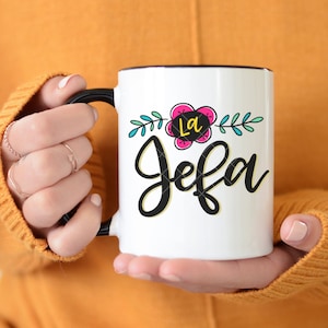 May include: White ceramic mug with a black handle and black rim. The mug has a floral design with a pink flower and green leaves. The text "La Jefa" is written in black cursive lettering.