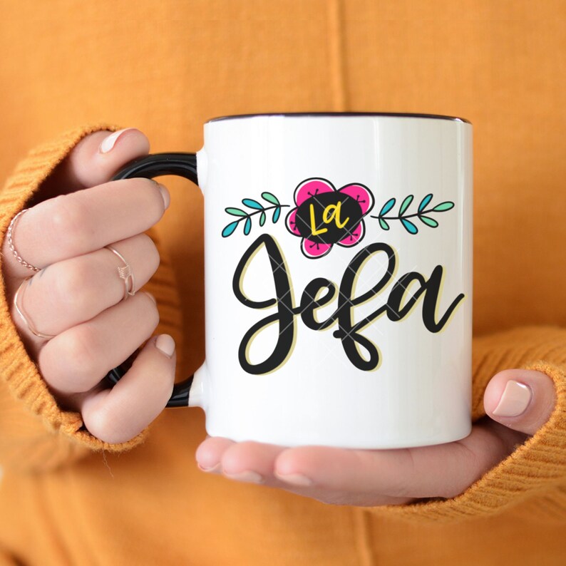May include: White ceramic mug with a black handle and black rim. The mug has a floral design with a pink flower and green leaves. The text "La Jefa" is written in black cursive lettering.