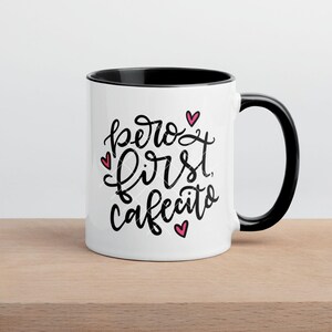 May include: White ceramic mug with a black rim and handle. The mug has a black handwritten saying "pero first cafecito" with three pink hearts.