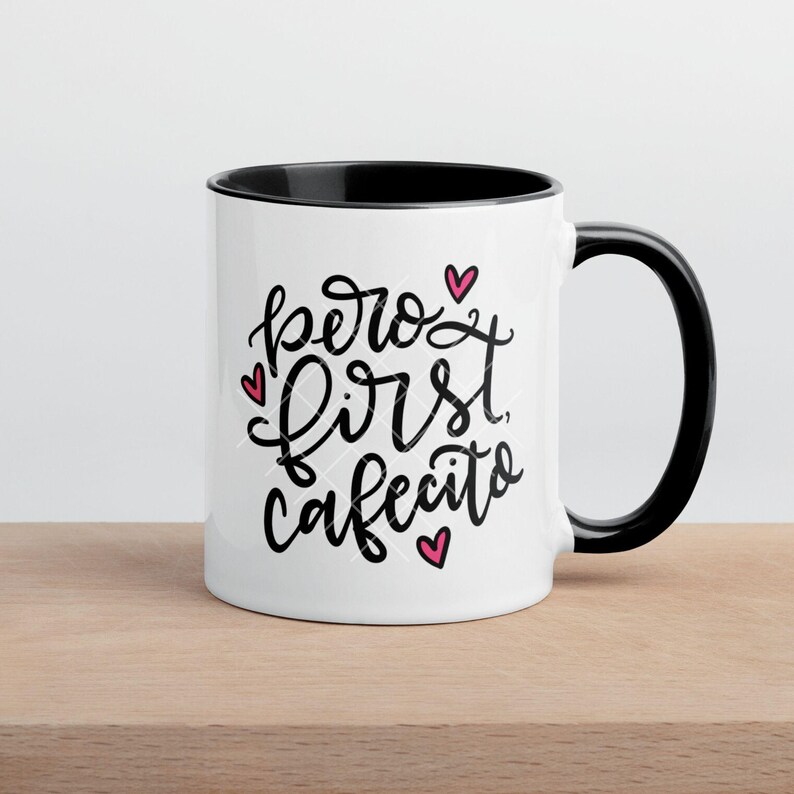 May include: White ceramic mug with a black rim and handle. The mug has a black handwritten saying "pero first cafecito" with three pink hearts.