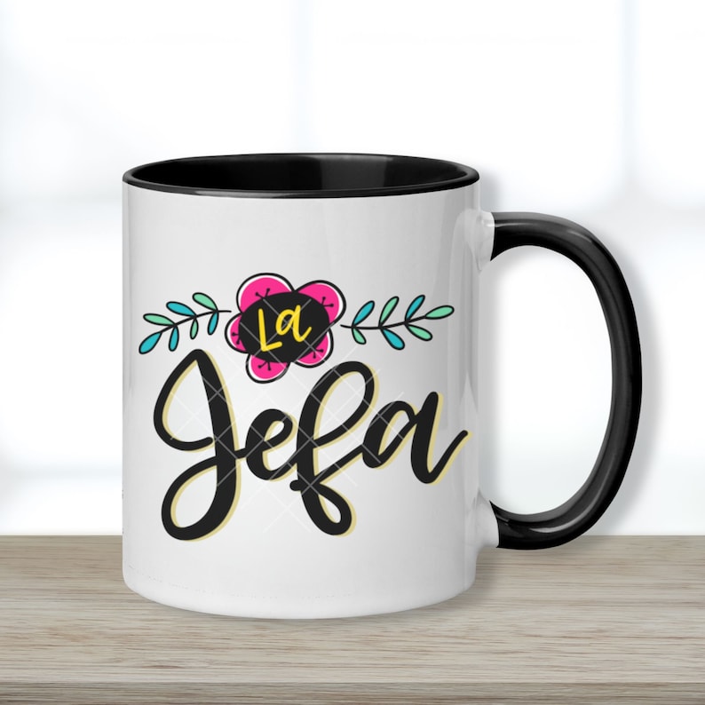 May include: White ceramic mug with a black rim and handle. The mug features a colorful floral design with the text "La Jefa" in black and yellow lettering.