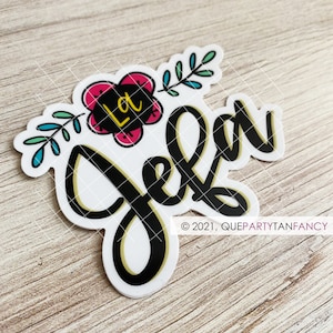 May include: A white sticker with the word "Jela" in a black script font with a gold outline. A pink flower with the letters "La" in yellow and green leaves are above the word. The sticker is on a light brown wooden surface.