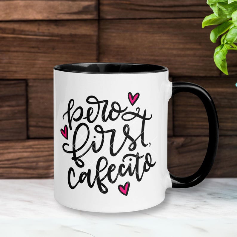 May include: White ceramic mug with a black rim and handle. The mug has a pink heart design and the text "pero first cafecito" in black.